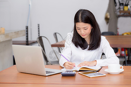 亚洲女商务人士用笔记本电脑在桌上的笔记本上写字 g商业女士笔记人士商务学生键盘计算器金融数据图片