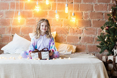 穿着睡衣的可爱女孩坐在床上 打开礼物和新年花冠图片