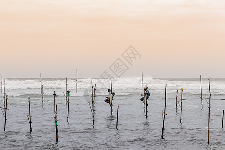 在日落的傍晚 几个高跷渔民坐在他们的杆子上 手里拿着木制钓鱼竿 海浪在他们身后撞击 背景是橙黄色的天空传统鱼人钓鱼文化热带旅行地图片