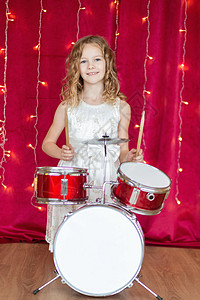 鼓凳笑笑的小女孩在红背景的鼓上打鼓 与新年花冠微笑女孩假期裙子花环礼物盒魔法孩子工作室盒子背景