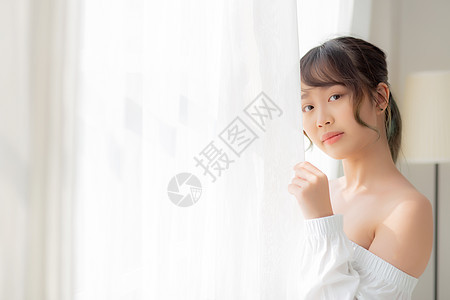 美丽的年轻亚洲女人的肖像 站在窗前房子房间休息阳光女性酒店公寓百叶窗幸福女士图片