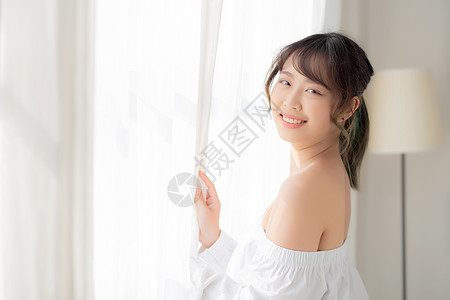 美丽的年轻亚洲女人的肖像 站在窗前公寓女孩睡衣女性酒店阳光房子快乐百叶窗休息图片