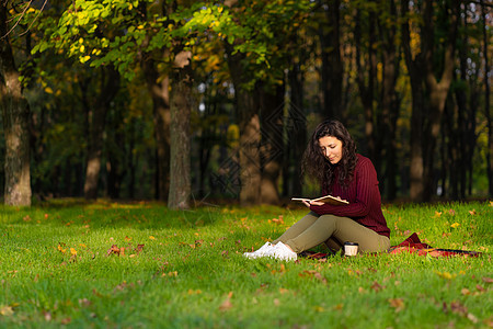 一个可爱的女孩读了一本书 喝着咖啡 在绿草坪上假期阳光树叶叶子公园学习杯子文学女性森林图片