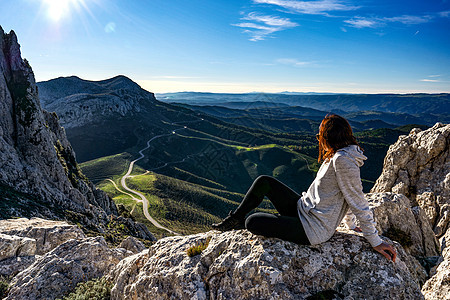美丽的年轻白人女性坐在一块白色的大石头上 俯视着美丽的全景 那里有一条从山上爬到地平线的路 — 无忧无虑的独行女性旅行者在休息图片