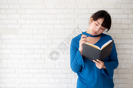 美丽的亚洲女性 微笑着站立思考和笔记本写在家中混凝土水泥白色背景上 在书籍 教育和生活方式概念上做女孩功课青少年家庭作业商业写作图片
