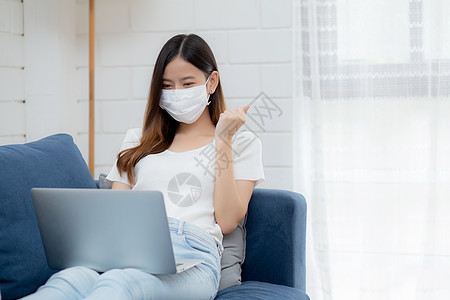 戴着面罩的年轻亚洲女商务人士从家里在笔记本电脑上在线工作 到客厅在互联网上取得成功 戴着医用口罩的女孩和 covid19 隔离区图片