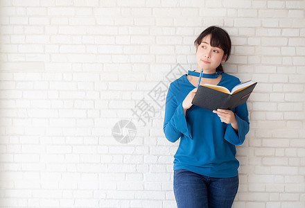 美丽的亚洲女性 微笑着站立思考和笔记本写在家中混凝土水泥白色背景上 在书籍 教育和生活方式概念上做女孩功课幸福成功工作考试学生商图片