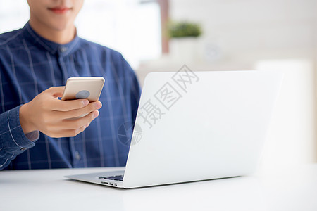 在家里的办公桌上工作笔记本电脑和在互联网上在线阅读智能手机的年轻人的特写手 自由职业者男性使用具有社交媒体 商业和通信概念的电话图片