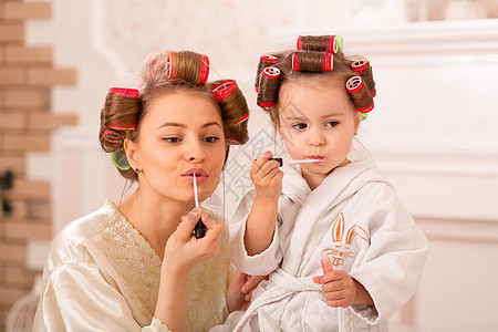 可爱的小女孩和她妈妈在卷发器上化妆 妈妈教女儿使用化妆品 美容日 女孩就是这样的女孩发型女士母性魅力闲暇嘴唇乐趣口红童年浴衣图片