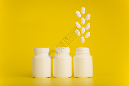 三个白色药瓶 最后一个打开 药丸在黄色背景下从里面跳出来图片