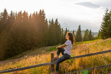 女孩坐在森林附近的高原田地的木栅栏上享受日落天空背包岩石女性乐趣裙子爬坡自由远足太阳图片