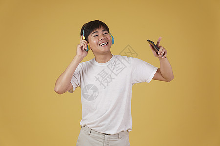 带着耳机的年轻亚洲人 听着智能手机上的音乐和在黄色背景下与世隔绝的舞蹈 笑声学生收音机韵律列表男性旋律工作室细胞电话乐趣图片
