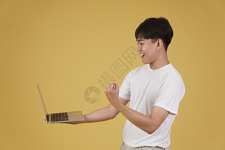 喜悦快乐的年轻亚洲男子穿着随意携带笔记本电脑 在黄色背景上孤立地做拳击赢家手势式动作技术男性商业互联网成人胜利商务优胜者工作人士图片