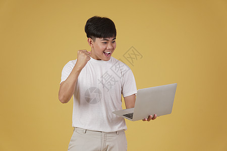 喜悦快乐的年轻亚洲男子穿着随意携带笔记本电脑 在黄色背景上孤立地做拳击赢家手势式动作胜利优胜者成人技术互联网男性学生男人人士工作图片