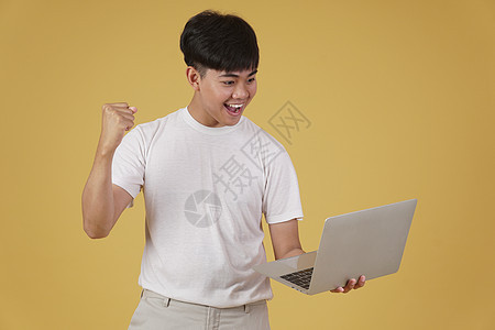 喜悦快乐的年轻亚洲男子穿着随意携带笔记本电脑 在黄色背景上孤立地做拳击赢家手势式动作优胜者人士工作商务技术互联网男人胜利学生工作图片