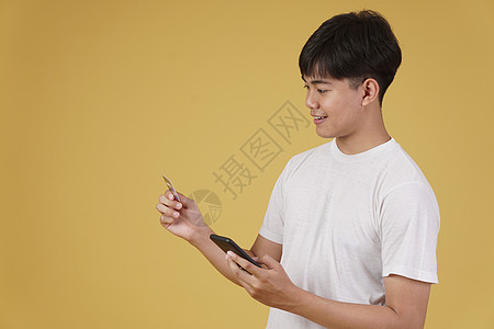 拥有智能手机和信用卡的快乐年轻亚洲男子肖像 用黄色背景与世隔绝的网上购物卡电话支付微笑工作室商业细胞男人开支享受购物者图片