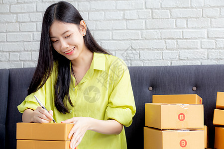 年轻的亚洲女性在家里的包裹箱上写地址 以便向客户 运输和物流 在线商家和卖家 企业主或中小企业 在线购物和电子商务订购送货单互联背景图片
