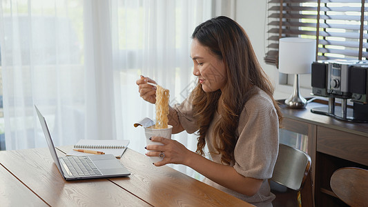 女商务人士在用笔记本电脑工作时吃即食面微笑人士女孩自由职业者桌子商务员工办公室食物女士图片