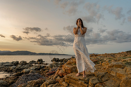 穿着白色长裙的沉思女人双手交叉放在脸前 闭上眼睛凝视着海上的日落 体验自然感觉良好并找到你的灵性的概念图片