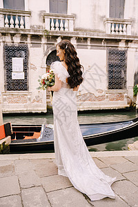 意大利威尼斯婚礼 一位穿着白色礼服的新娘 乘坐火车 手捧一束白色和红色的玫瑰 站在狭窄的威尼斯运河中停泊的贡多拉附近的码头上桉树图片