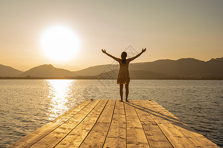 年轻女性穿着短裙赤脚在木制码头上露着双臂 观察湖面或海面山中太阳落山的景象;橙色老式摄影效果图片
