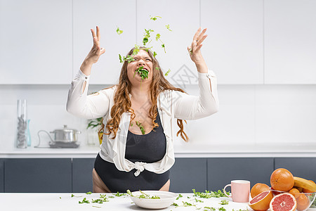 快乐的胖女孩把菠菜叶撒得满桌都是 超重的女孩快乐减肥吃新鲜的沙拉 曲线优美的身体 留着长长的金发的年轻女子 节食和营养概念体重秤图片