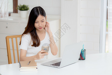 年轻的亚洲女性在家里的办公桌上工作笔记本电脑并在互联网上在线阅读智能手机 自由职业者女孩微笑着使用带有社交媒体 商业和通信概念的图片