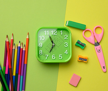 方形绿色时钟和绿色背景上的一叠多色木铅笔 顶视图图片