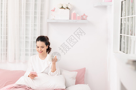 坐在房间床上喝咖啡和使用手机的年轻女学生图片