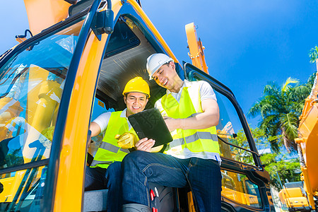 亚洲建筑司机与工程师蓝图讨论工业挖掘机头盔药片软垫工地机械黄色男人导师图片