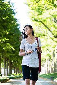 在公园中行走的清廉的年轻女子长廊太阳镜闲暇黑发耳环女性叶子手提包游客配件背景图片
