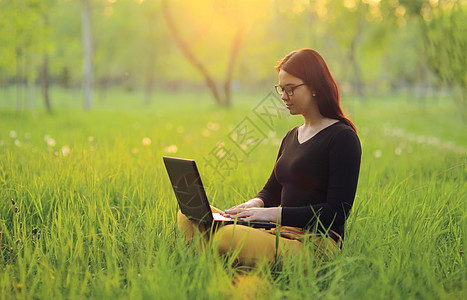 带着笔记本电脑的可爱笑笑女孩坐在公园绿草地上 作为自由职业者远程工作图片
