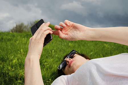 带着眼镜的女孩躺在绿草上 拿着智能手机成人电话女士花园自拍电子产品公园黑发阅读日光图片