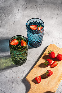 两个绿色和蓝色几何玻璃杯 配有淡水和草莓水果 在石头混凝土背景 角度视图的木切板附近贴着多彩色影子光线图片