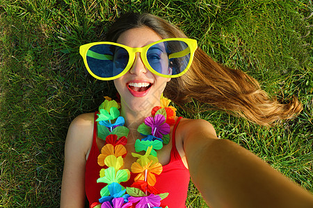 女孩在狂欢节派对后自拍 年轻女子的自拍 戴着滑稽的大太阳镜和躺在草地上的嘉年华花环图片