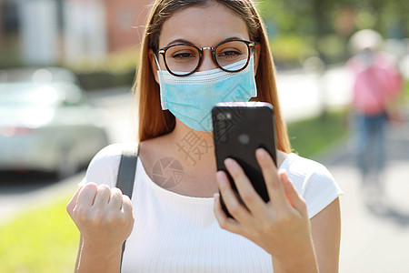 兴奋的年轻女商务人士 她带着外科面罩在手机上听到好消息 在街上用拳头庆祝一番图片