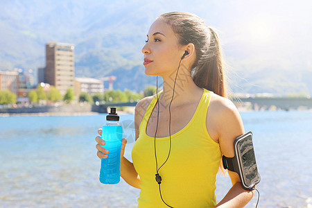 手持智能电话臂带的干渴女运动员拿着电水饮料和户外观望图片