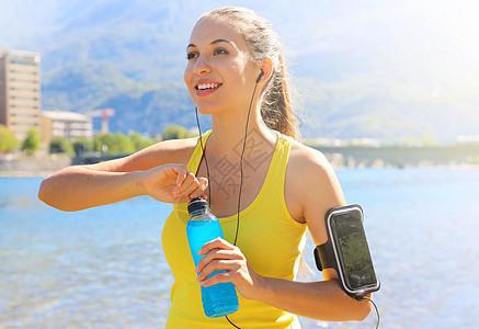 拥有智能手机开源饮料瓶的臂带 在日常训练后愉快地欢乐女性运动员在户外观望图片