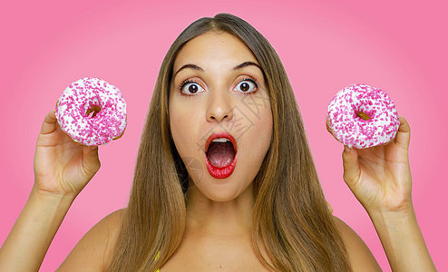 惊讶的年轻女人拿着两块甜甜圈 在她手上握着粉红色背景图片