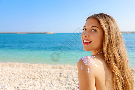 肩上有太阳形状的年轻女子 美丽快乐可爱的女人在她的肩膀上涂上防晒霜 背景是海滩 复制空间图片