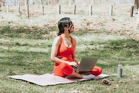 身穿健身服的年轻女性在公园的笔记本电脑上放松 在带复制空间的瑜伽垫上图片