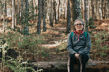 坐在树干旁边的老女人 在秋天在森林里微笑着拍镜头 带着复制空间 健康漫步于慢跑的概念图片