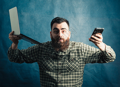 有留胡子概念的年轻时装男青年 用笔记本电脑和电话在蓝色背景上工作 要求照相机复制空间图片