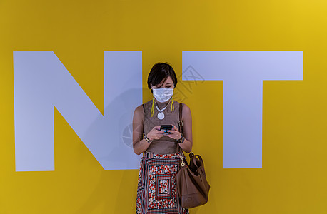 一名年轻时身戴面罩 使用智能电话打短信的年青妇女站在N字母和T字母中黄色墙背景前 (笑声)图片