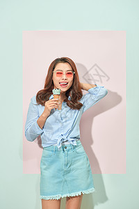 高兴的是 带着露齿微笑的可爱亚洲年轻女性看着镜头 拿着美味的冰淇淋 站在浅粉色背景中 身着旅游装 戴着眼镜黑发奶油白色甜点锥体乐图片
