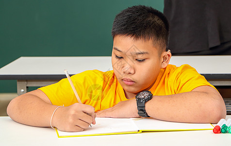 亚洲残疾男孩或穿着黄色衬衫的自闭症儿童 手持铅笔 学习在教室桌上集中阅读和写字 校对 Soup图片