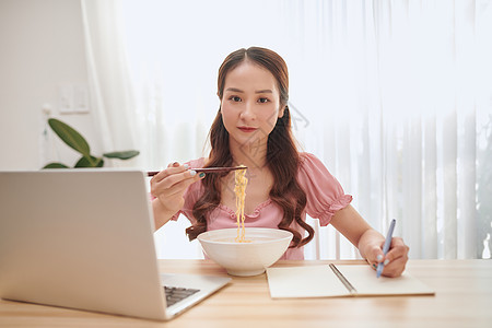 年轻亚洲女人吃面条 在家用笔记本电脑工作办公室长发写作快乐喜悦女士微笑商业牙齿筷子图片