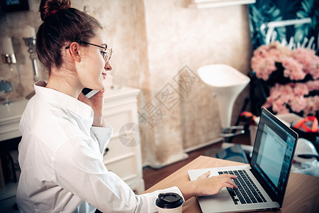 从家庭生活方式和在线商务工作 有魅力的女人手在家里工作时打电话给手机 商务自由职业者在她的家庭工作场所使用笔记本电脑 新常态女士图片