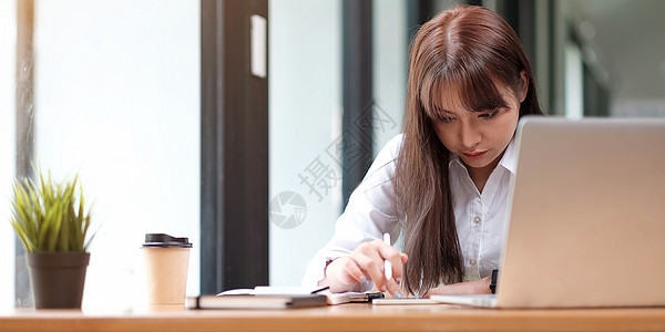 在线教育理念 严肃的女学生学习 观看网络研讨会 在笔记本上做笔记 坐在办公桌前 在家里或办公室用笔记本电脑并在商业规划师中写作的图片