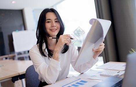 美丽的亚洲女商务人士拿着文书 铅笔和微笑看着相机 在明亮的现代办公室工作时笑着看镜头图片
